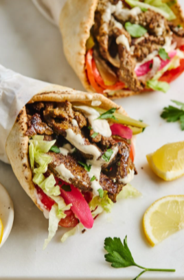 Homemade beef shawarma being wrapped in soft pita bread with lettuce, tomatoes, and garlic sauce.